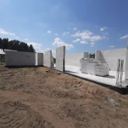 Wznoszone ściany z białych bloczków betonu komórkowego na fundamencie z widocznymi prętami zbrojeniowymi, na tle piaszczystego terenu budowy i błękitnego nieba.