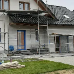 Dom jednorodzinny w trakcie prac elewacyjnych: ściany pokryte szarym tynkiem, okna i drzwi zabezpieczone folią, rusztowania wokół budynku, widoczne panele słoneczne na dachu.