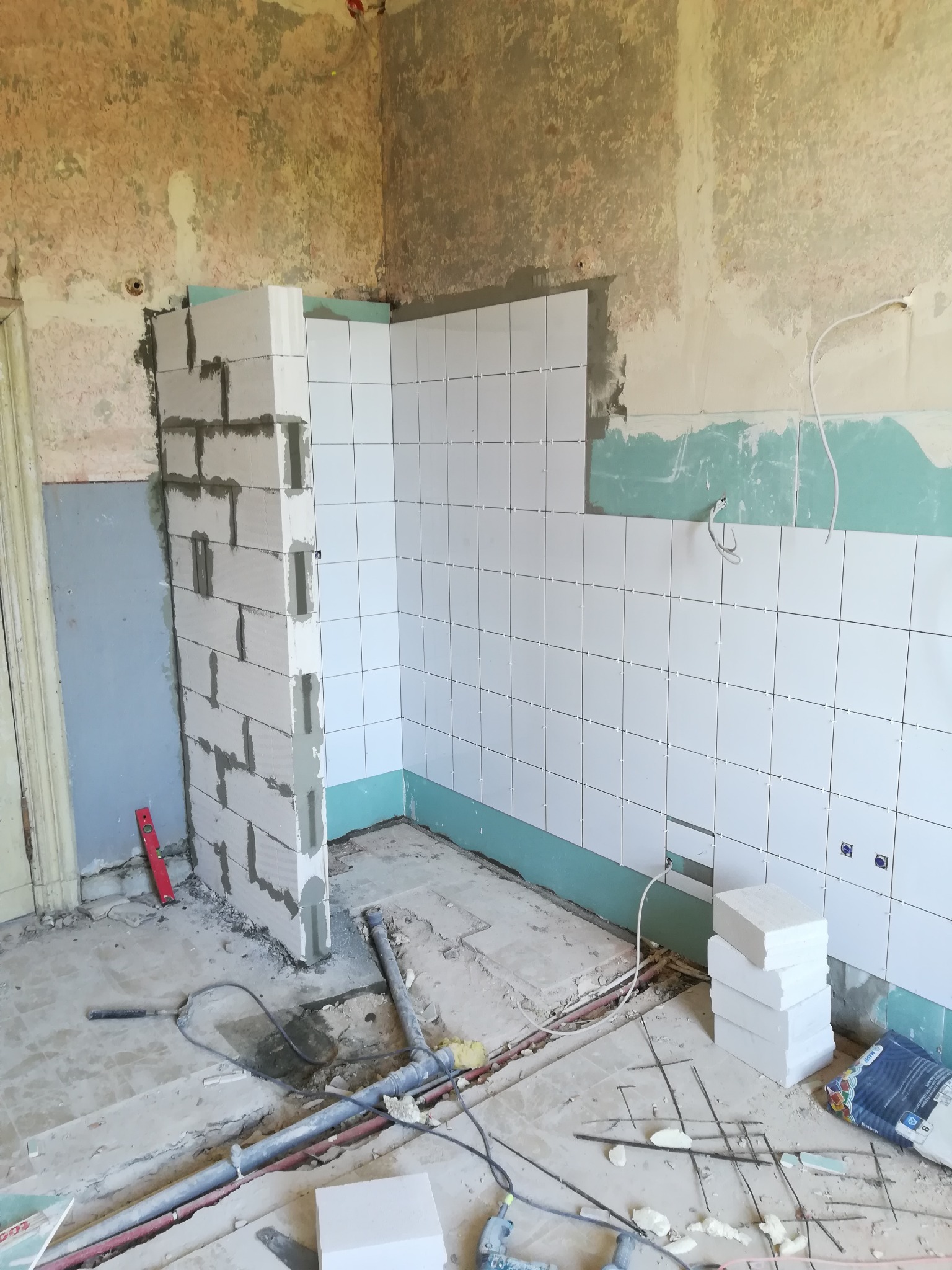 Wnętrze łazienki w trakcie układania białych płytek ściennych, widoczna ściana z bloczków betonowych, poziomica i instalacje podłogowe.