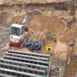 Głęboki wykop z widocznymi warstwami ziemi, mini koparka Takeuchi z przodu, zagęszczarka w wykopie, szalunki fundamentowe ułożone obok, zbrojenie i rury kanalizacyjne w tle.