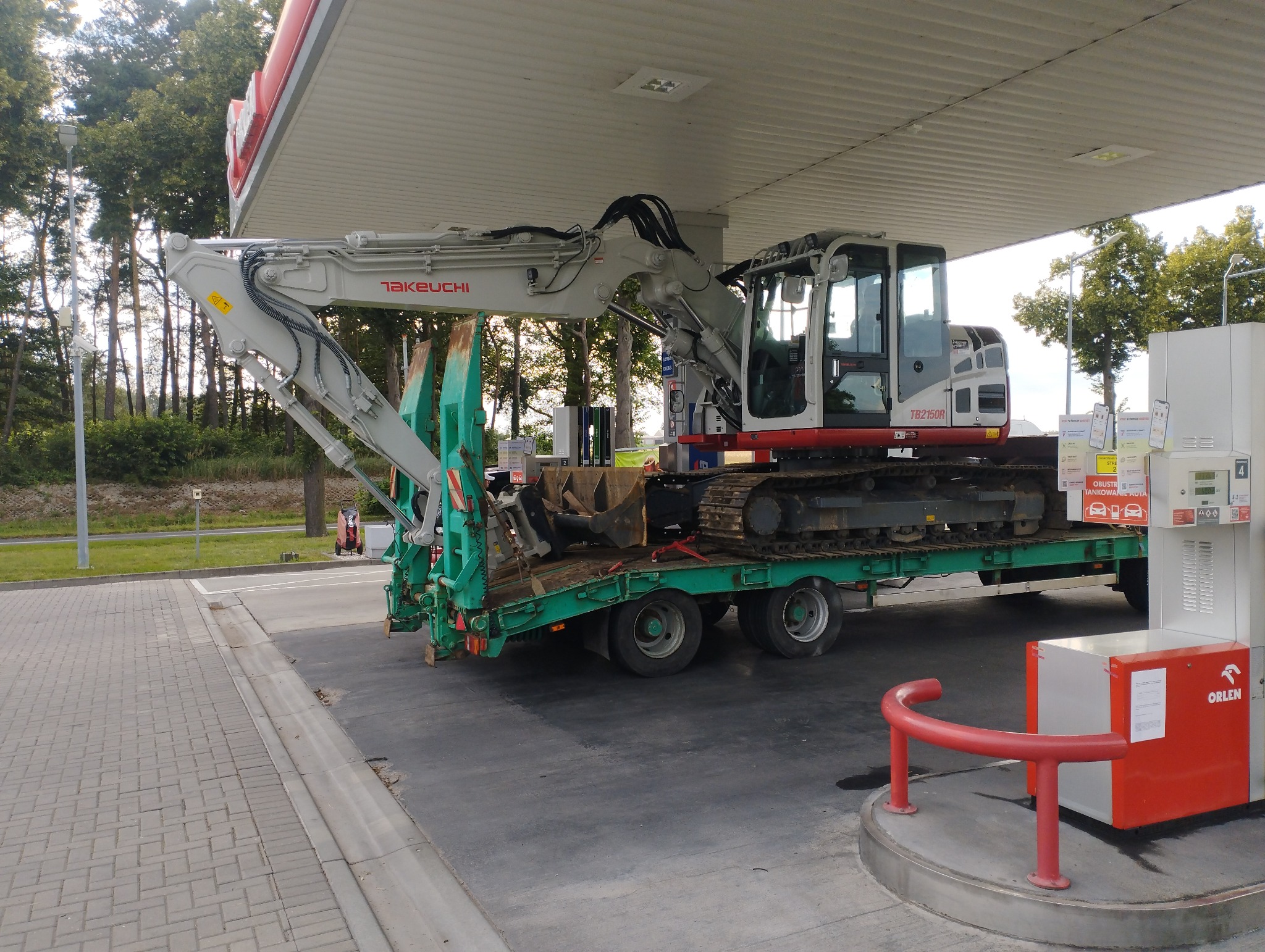 Koparka Takeuchi TB2150R na zielonej przyczepie zaparkowana pod wiatą stacji benzynowej Orlen, widoczne drzewa w tle.