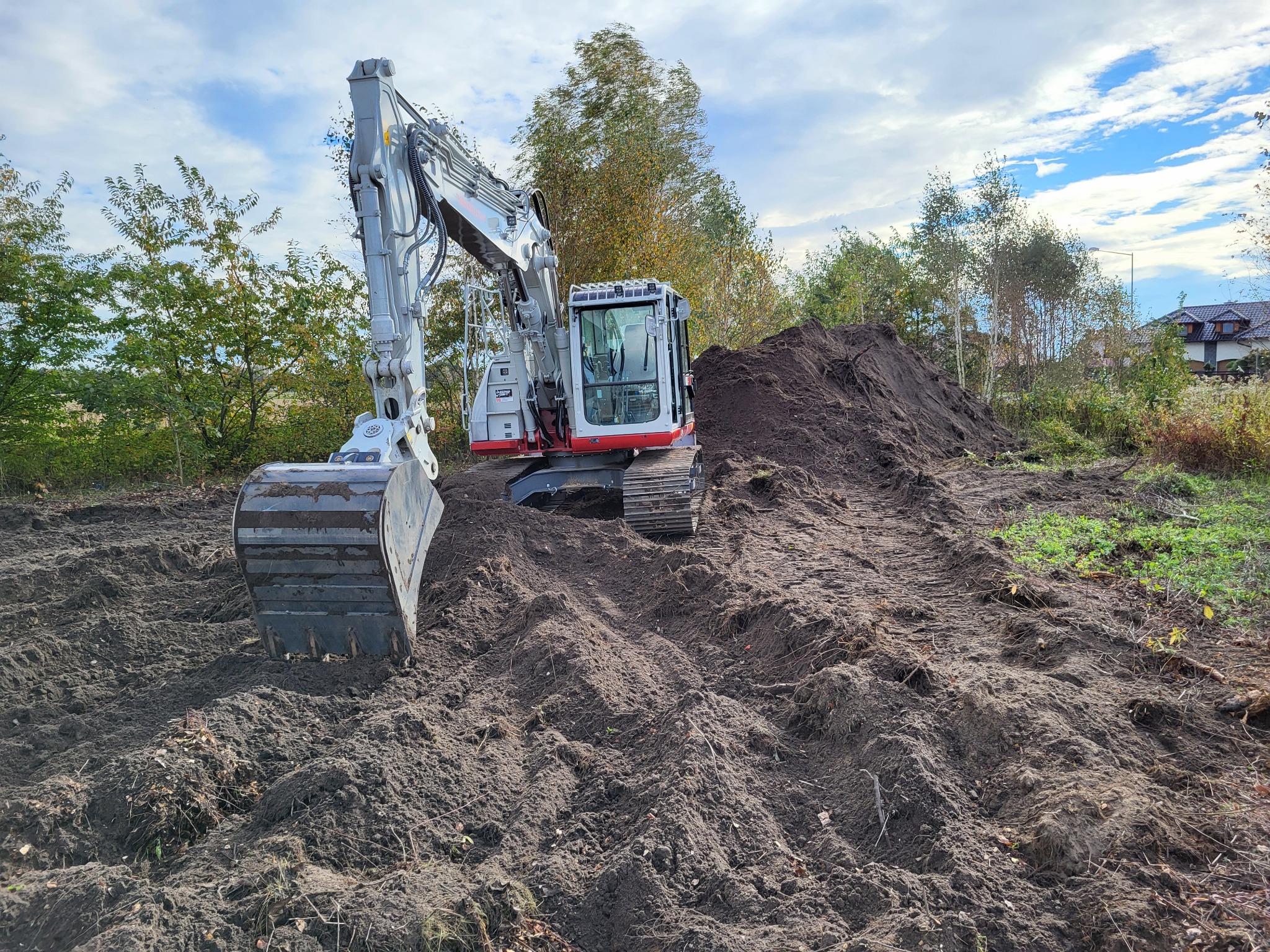 Biało-szara koparka na gąsienicach, z łyżką uniesioną do góry, stoi na nierównym terenie z pryzmą ziemi w tle, widoczne ślady gąsienic, drzewa i dom w oddali.
