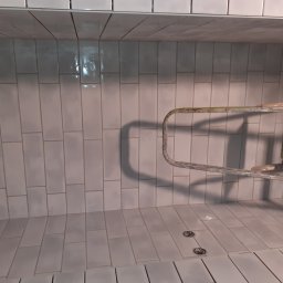 MAG-BUD Firma remontowo budowlana - Świeżo ułożone, pionowe, białe płytki w kształcie cegiełek, z widoczną, metalową drabinką na tle, oraz dwoma okrągłymi odpływami w podłodze.