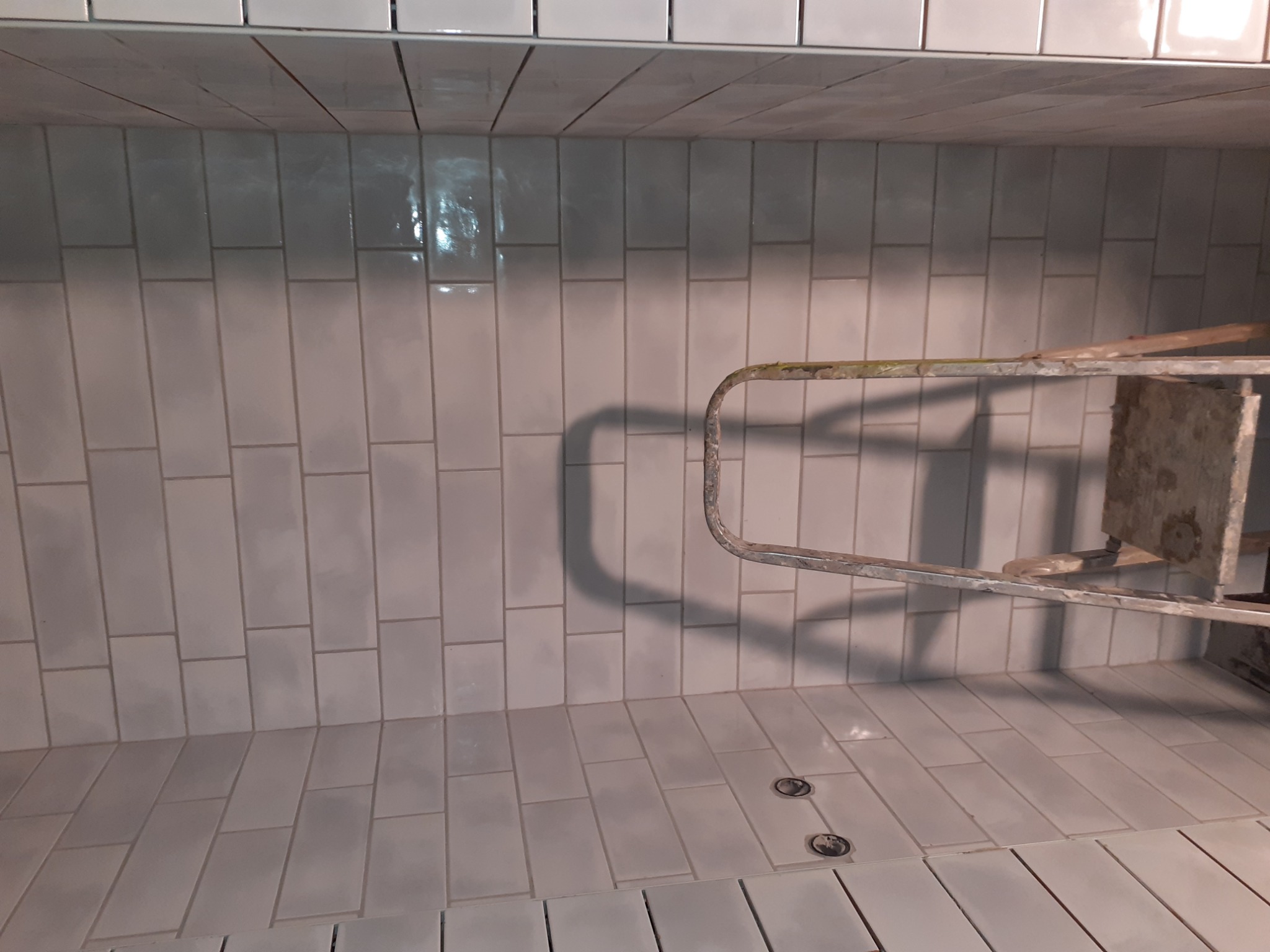 Świeżo ułożone, pionowe, białe płytki w kształcie cegiełek, z widoczną, metalową drabinką na tle, oraz dwoma okrągłymi odpływami w podłodze.