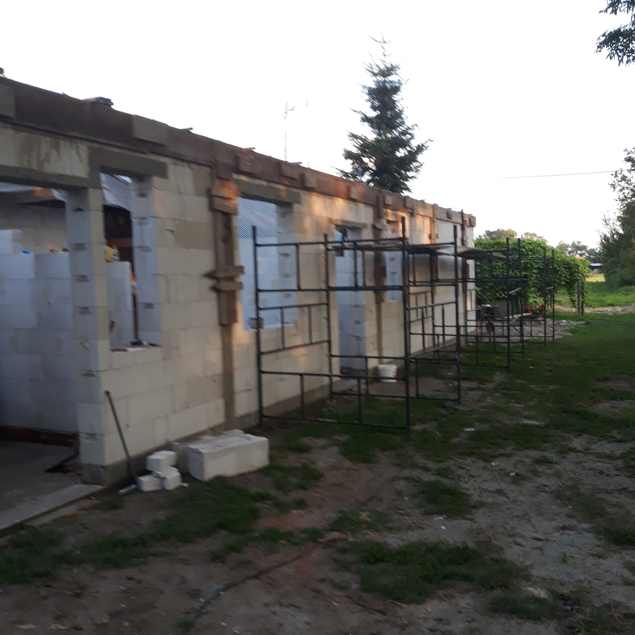 Surowy stan budynku w trakcie budowy z białych bloczków, widoczne rusztowania i elementy konstrukcyjne, otoczenie placu budowy.