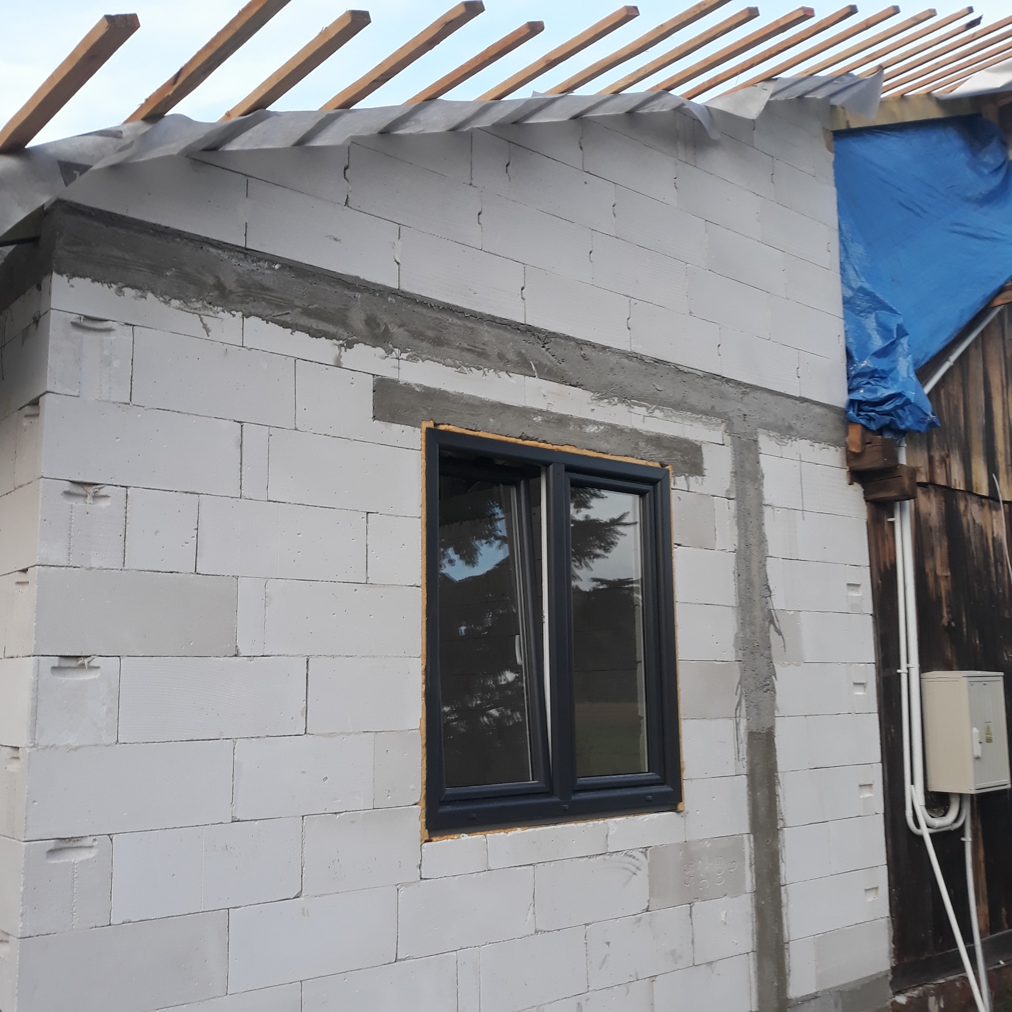 Ściana budynku w trakcie budowy z białych bloczków, z zamontowanym oknem z czarną ramą, widoczne drewniane elementy dachu i niebieska plandeka.