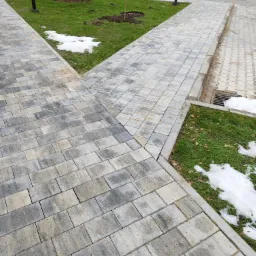 Szeroki kadr na skrzyżowanie chodników z kostki brukowej w odcieniach szarości i beżu, z fragmentami trawnika z resztkami śniegu, w tle zaparkowane samochody i przechodnie.