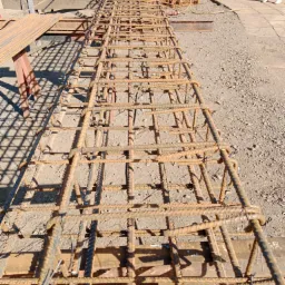 Szkielet z prętów zbrojeniowych, widok z góry, z detalami wiązań drutem i cieniem rzucanym na żwirowe podłoże.