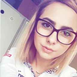 Portret kobiety w okularach o ciemnych oprawkach, z blond włosami do ramion i delikatnym uśmiechem, ubrana w białą koszulę z czarno-białą szachownicą przy kołnierzyku, sfotografowana...