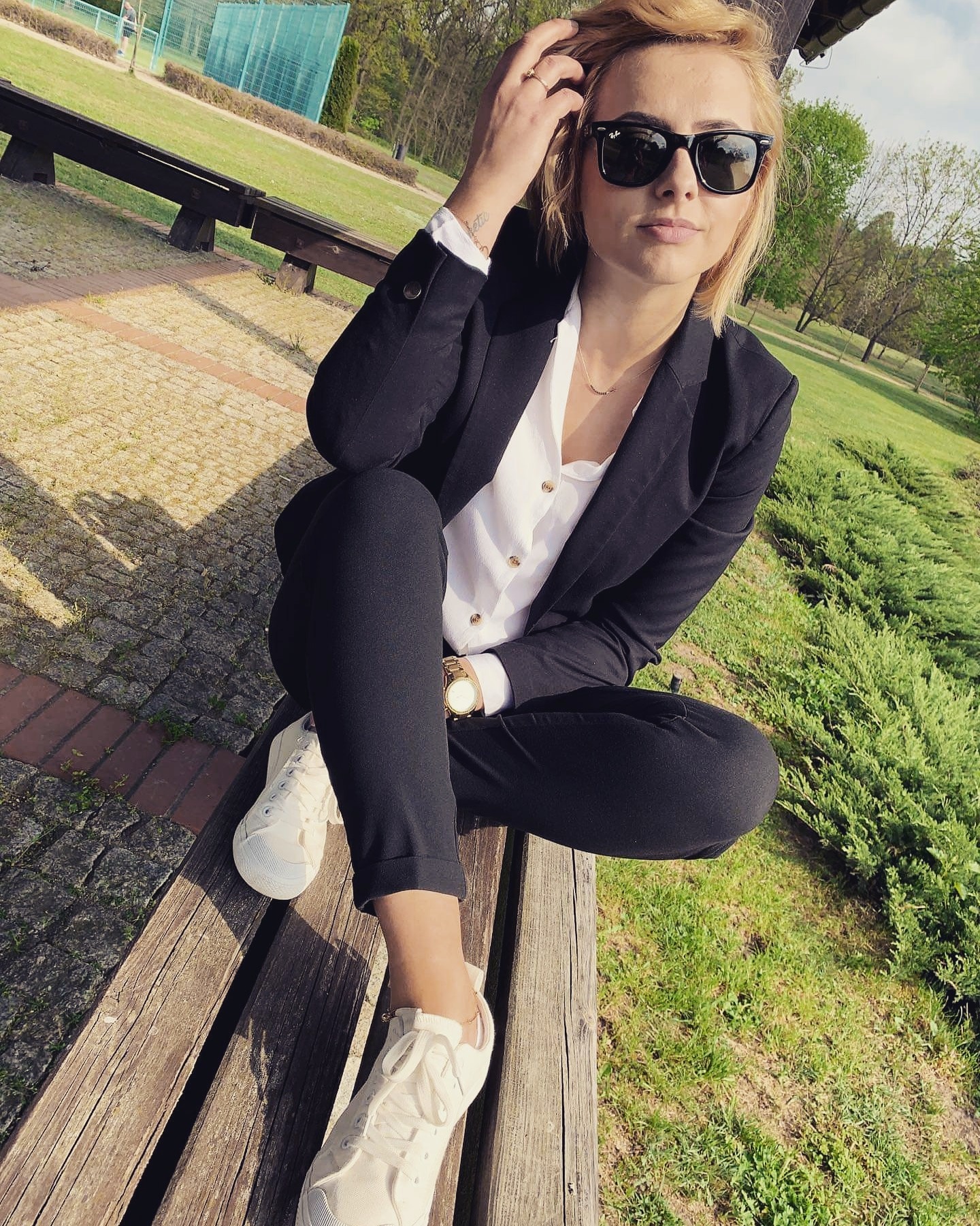 Portret kobiety w czarnym garniturze i białych trampkach, siedzącej na drewnianej ławce w parku w słoneczny dzień, z okularami przeciwsłonecznymi.