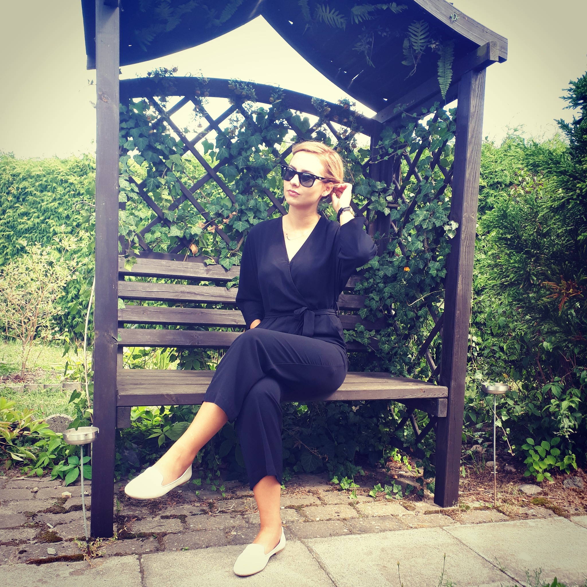 Kobieta w czarnym kombinezonie i okularach przeciwsłonecznych siedzi na drewnianej ławce pod zadaszeniem oplecionym bluszczem, w tle zielony ogród.