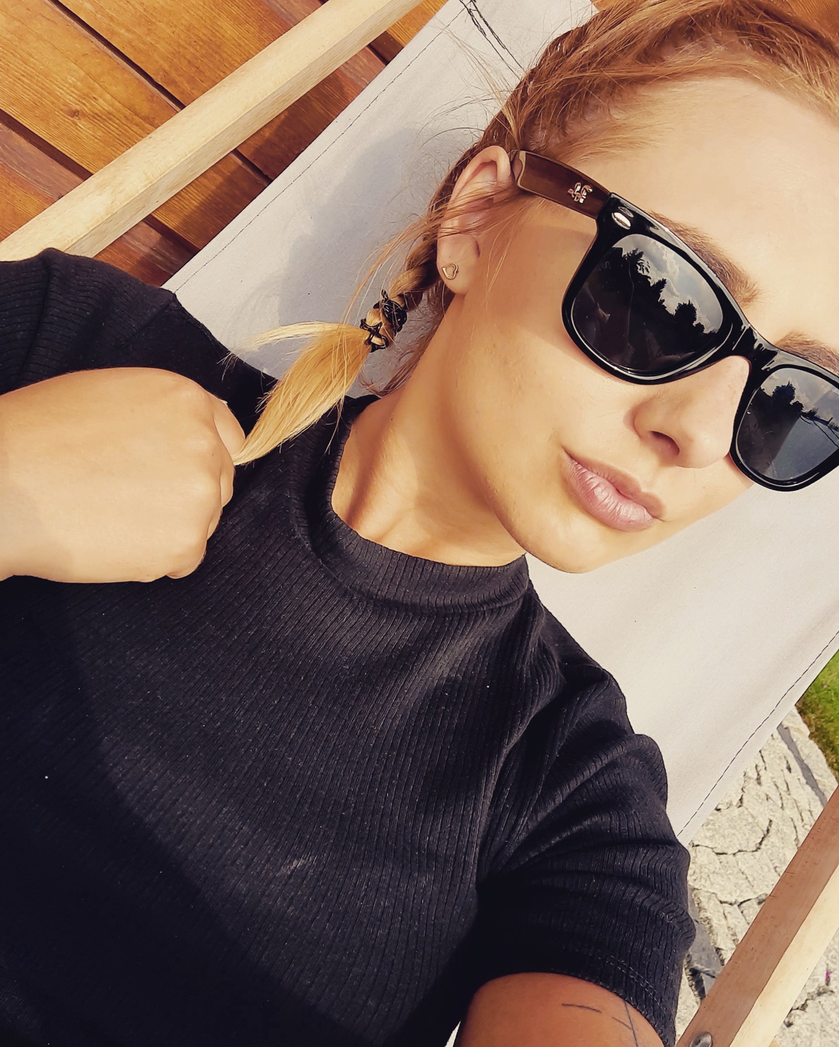 Portret kobiety w czarnej bluzce i okularach przeciwsłonecznych, leżącej na leżaku z plecionym warkoczem i złotymi kolczykami w kształcie serca.