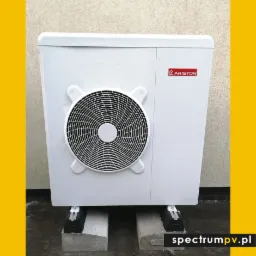 Biała jednostka zewnętrzna z logo Ariston, umieszczona na betonowych blokach z amortyzatorami, na tle jasnej ściany.