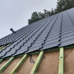 Montaż szarej blachodachówki modułowej na łatach z użyciem wkrętarki akumulatorowej, widoczny kominek wentylacyjny i membrana dachowa.