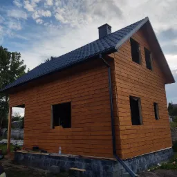Surowa drewniana konstrukcja domu z ciemnym dachem z blachodachówki, widoczne niezabezpieczone otwory okienne i brakujące elementy wykończeniowe, częściowo ukończona elewacja, na fundamencie...
