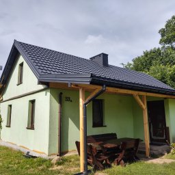 MAR-DACH Mariusz Boruc - Jasnozielony dom z ciemnym dachem z blachy trapezowej i drewnianą pergolą nad wejściem. Widoczny numer domu '33a'.