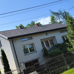 MAR-DACH Mariusz Boruc - Dwupiętrowy dom z szarą elewacją i ciemnym dachem, widoczny zza metalowego ogrodzenia. Uwagę zwraca polska flaga w oknie na piętrze i iglak przy ścianie.