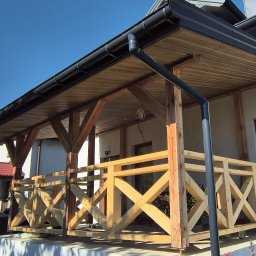 MAR-DACH Mariusz Boruc - Zadaszony taras z drewnianą balustradą w surowym stanie, widok z dołu. Czarna rynna i fragment elewacji domu w tle, błękitne niebo.