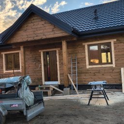 MAR-DACH Mariusz Boruc - Drewniany dom szkieletowy z ciemnym dachem z blachodachówki, widoczny z zewnątrz, z elementami budowy i narzędziami na pierwszym planie. Widoczne okna i drzwi.