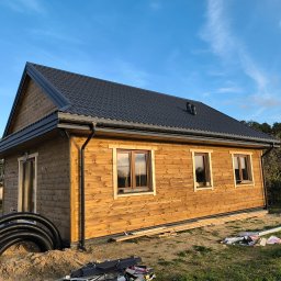 MAR-DACH Mariusz Boruc - Drewniany dom szkieletowy z ciemnym dachem i widocznymi oknami, otoczony zielenią i elementami budowlanymi. Ujęcie w słoneczny dzień.