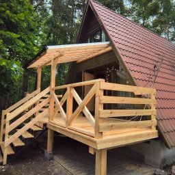 MAR-DACH Mariusz Boruc - Nowy drewniany taras z zadaszeniem przy domu z czerwoną dachówką, otoczony zielenią. Widoczne schody i balustrady. Świeże drewno, naturalne otoczenie.