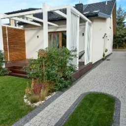 Nowoczesna, biała konstrukcja z drewnianym panelem i tarasem przylegająca do domu z brukowaną ścieżką i zadbanym trawnikiem.