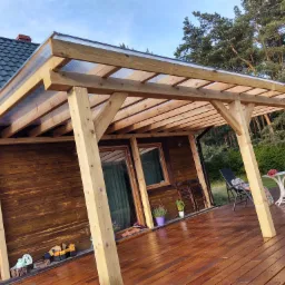 Zadaszony taras z drewnianą podłogą przy drewnianym domu, z meblami ogrodowymi i roślinami w doniczkach, widok na ogród i las w tle.