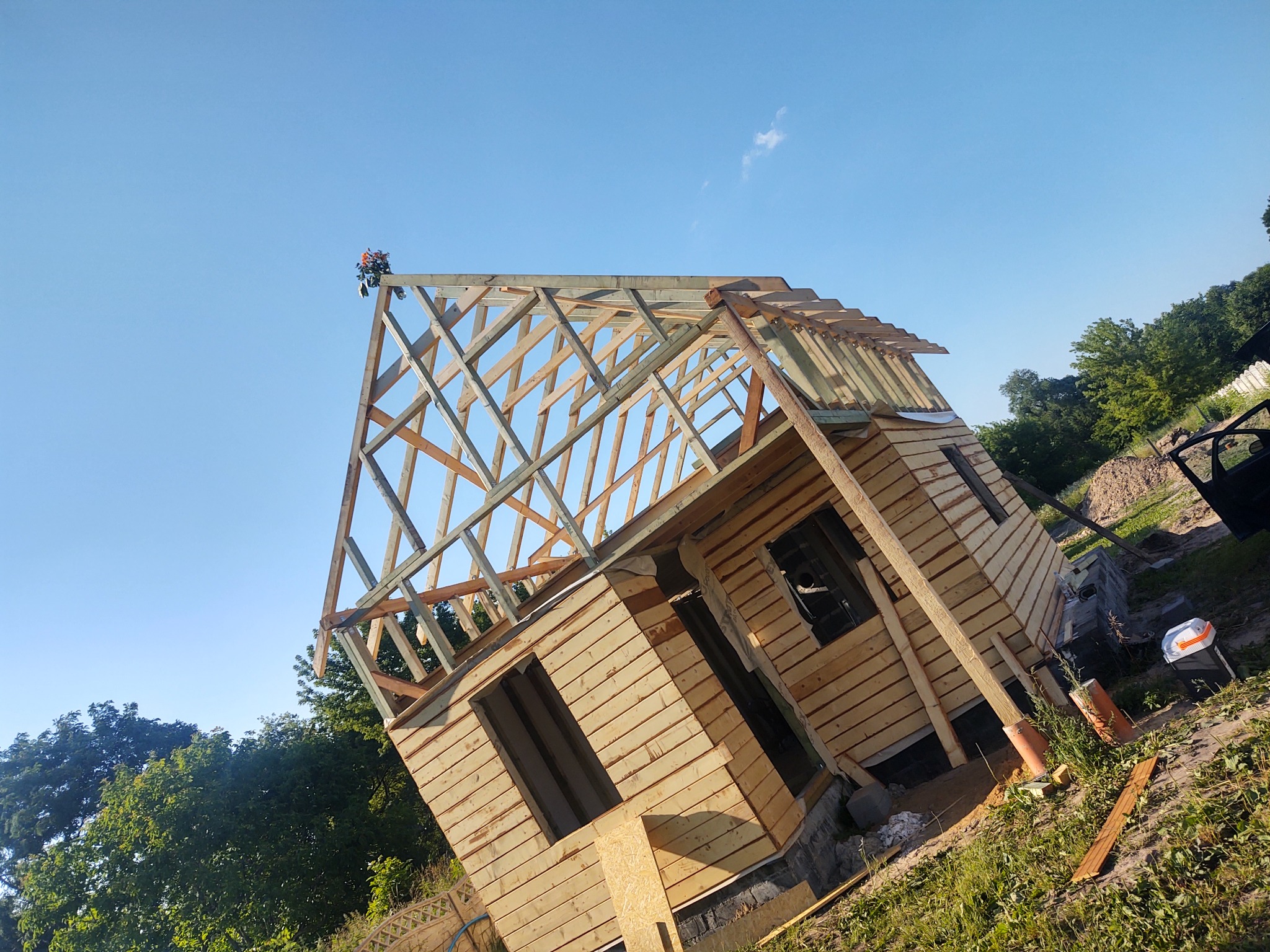 Budowa małego drewnianego domu z widoczną konstrukcją szkieletową dachu, na tle błękitnego nieba i zieleni drzew