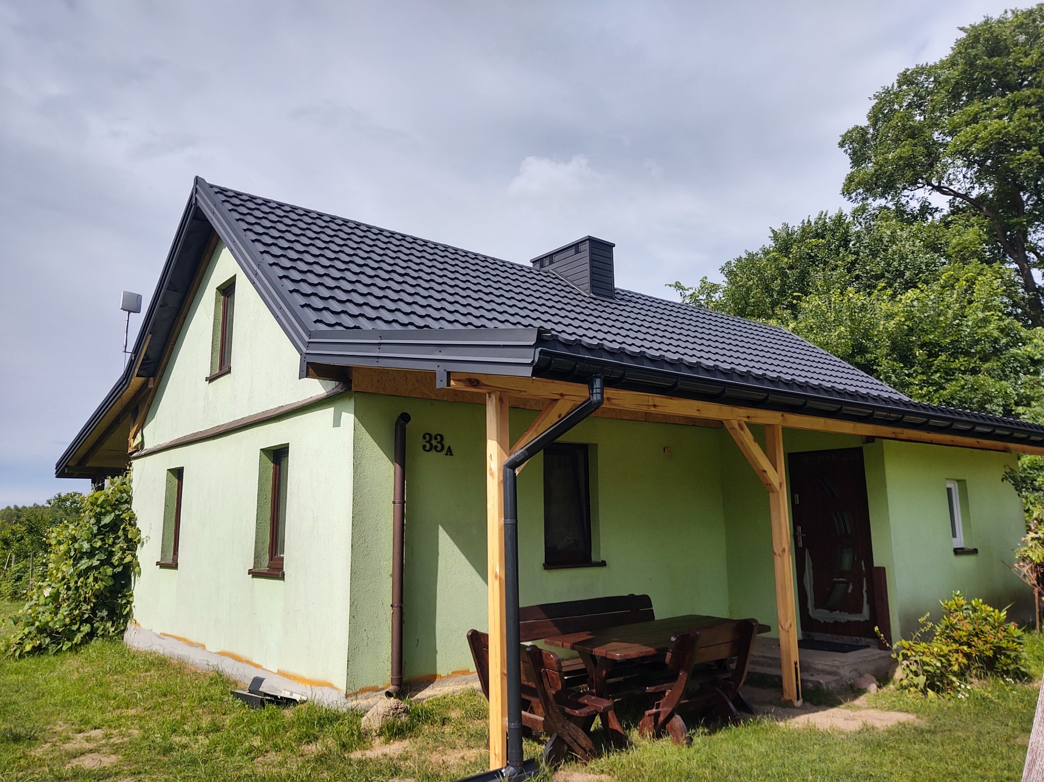 Jasnozielony dom z ciemnym dachem z blachy trapezowej i drewnianą pergolą nad wejściem. Widoczny numer domu '33a'.