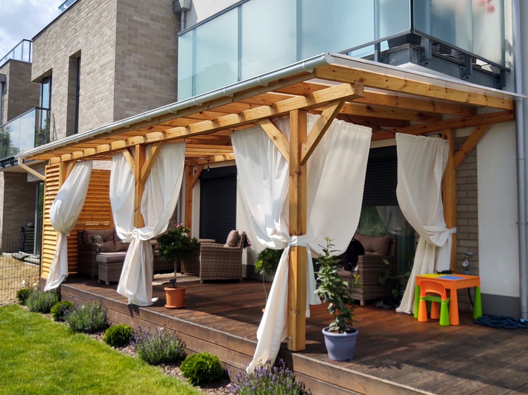 Drewniana pergola z białymi zasłonami na tarasie, przytulne meble ogrodowe, rośliny w donicach i mały stolik dla dzieci. Nowoczesny dom w tle.