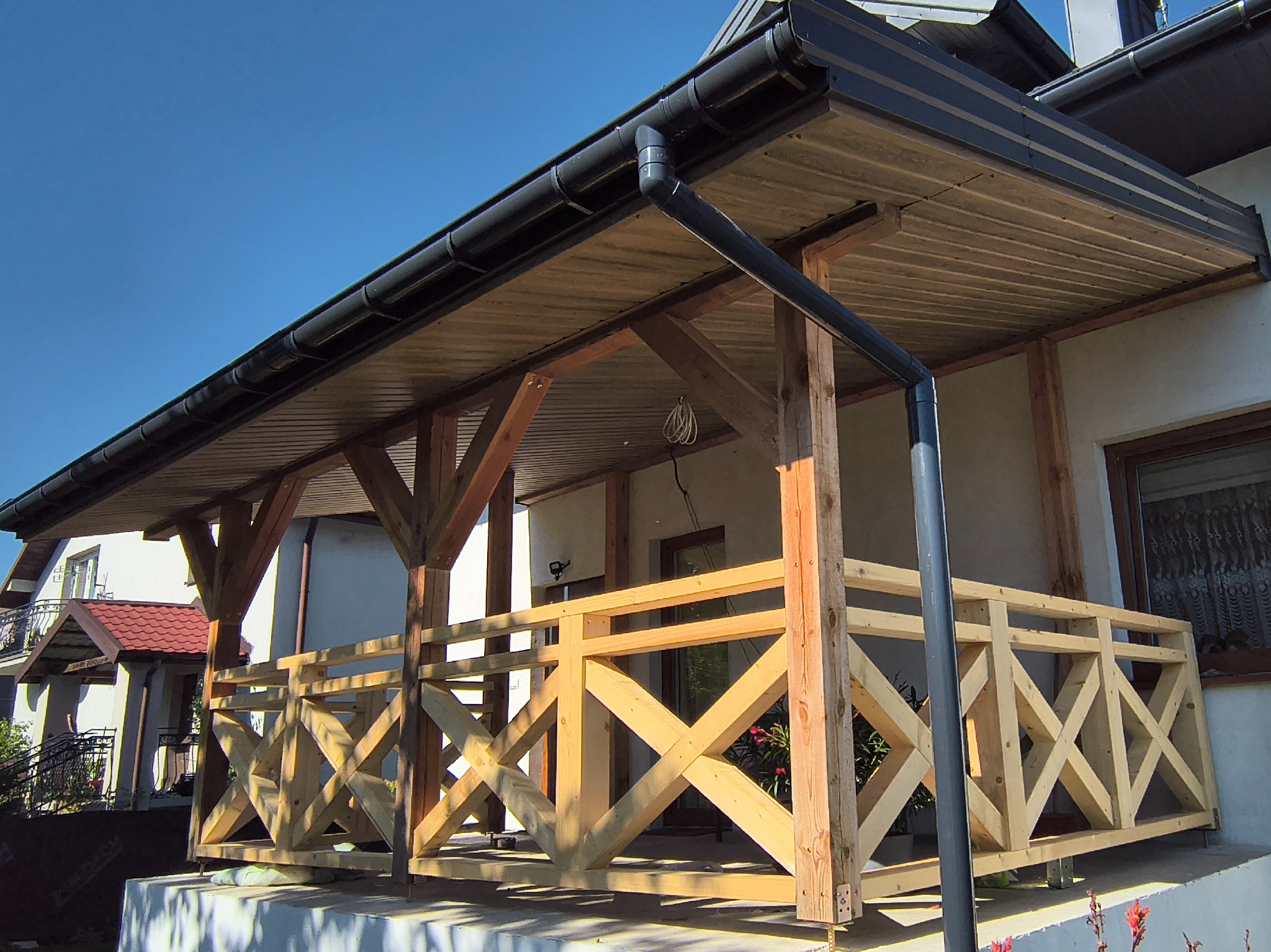 Zadaszony taras z drewnianą balustradą w surowym stanie, widok z dołu. Czarna rynna i fragment elewacji domu w tle, błękitne niebo.