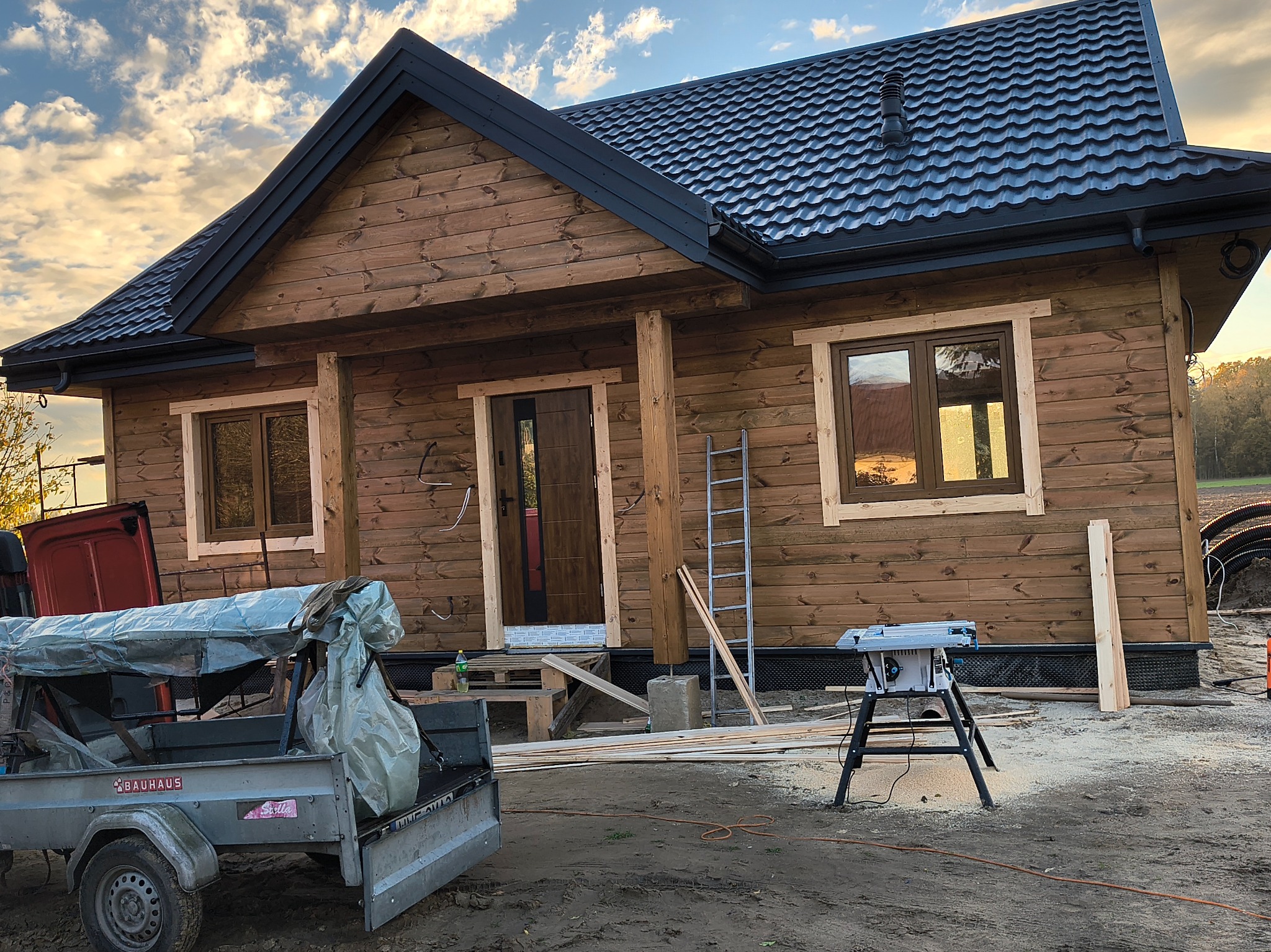 Drewniany dom szkieletowy z ciemnym dachem z blachodachówki, widoczny z zewnątrz, z elementami budowy i narzędziami na pierwszym planie. Widoczne okna i drzwi.