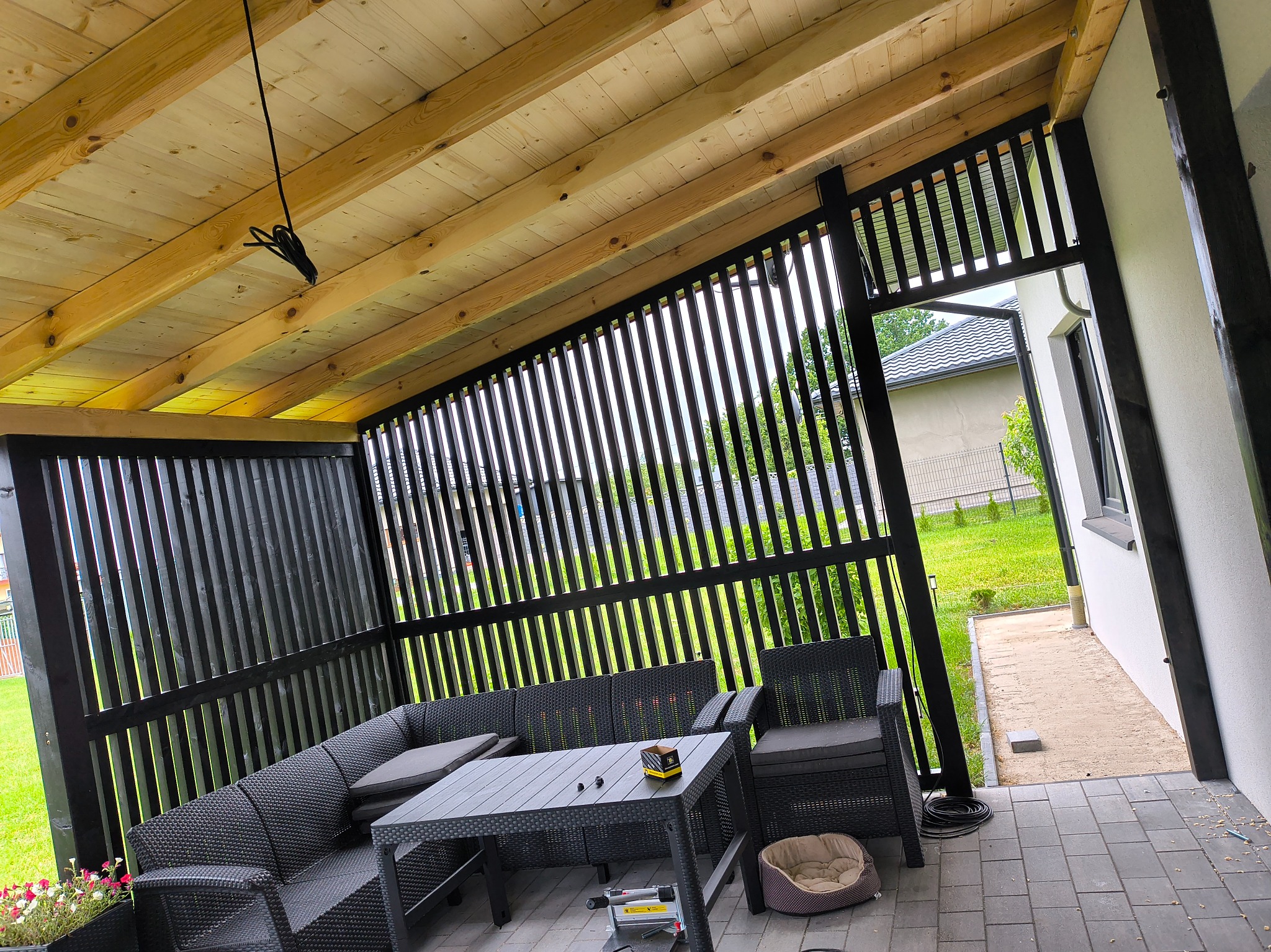 Zadaszona pergola z drewnianym sufitem i czarnymi, pionowymi panelami. Wewnątrz meble ogrodowe z rattanu i stolik. Widok na ogród i dom z zewnątrz.