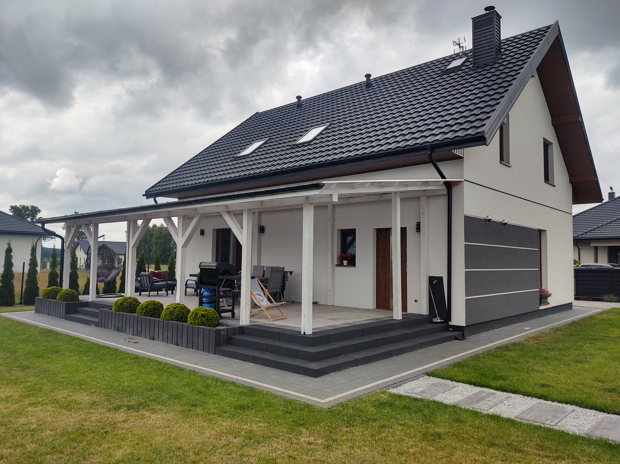 Nowoczesny dom z ciemnym dachem i jasną elewacją. Zadaszony taras z białymi kolumnami, schody z kostki, zadbany trawnik i dekoracyjne krzewy.