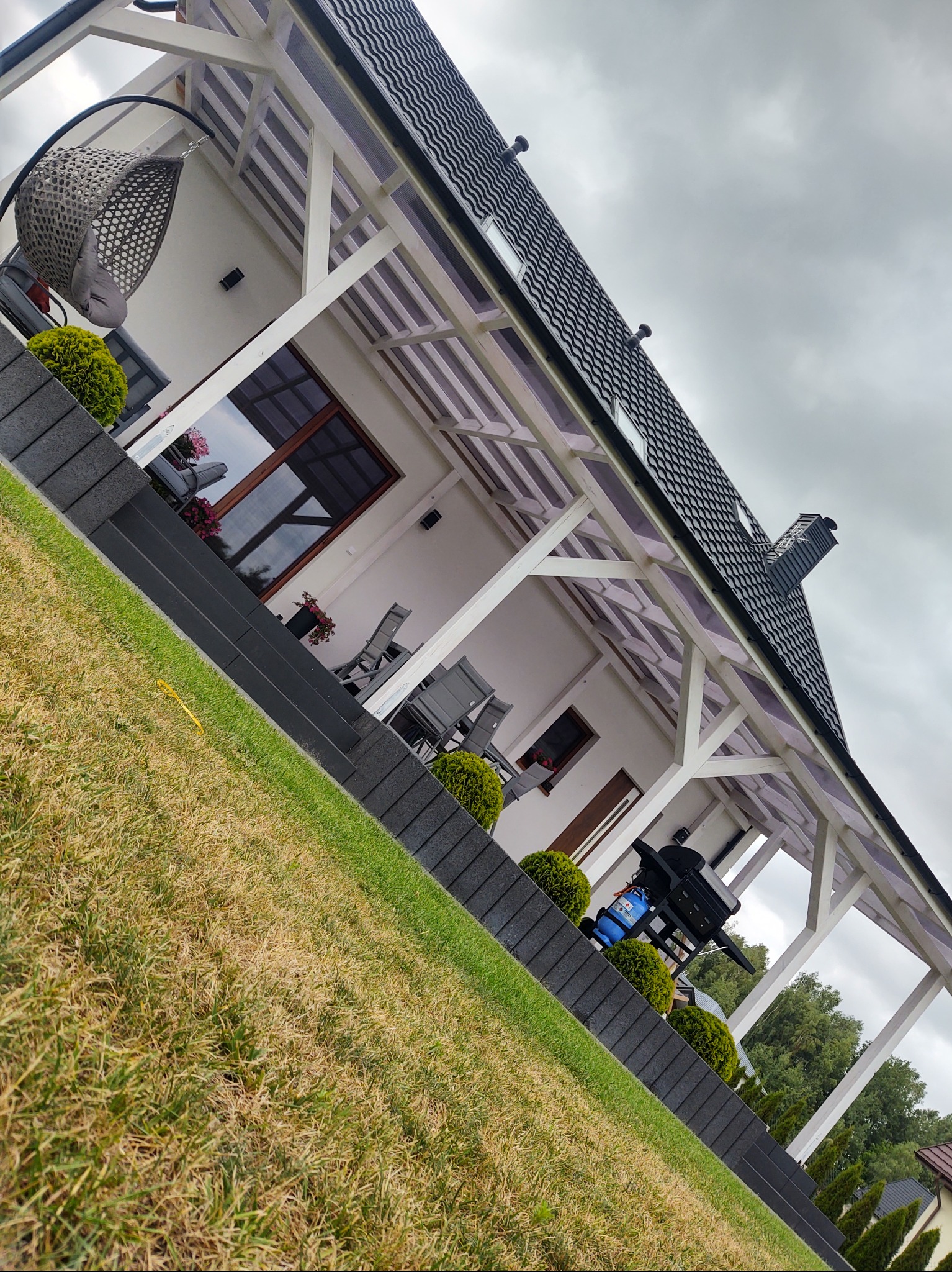 Biała pergola z przezroczystym zadaszeniem przy domu z ciemnym dachem. Na pierwszym planie trawnik i schody z kostki. W tle wiszące krzesło i meble ogrodowe.