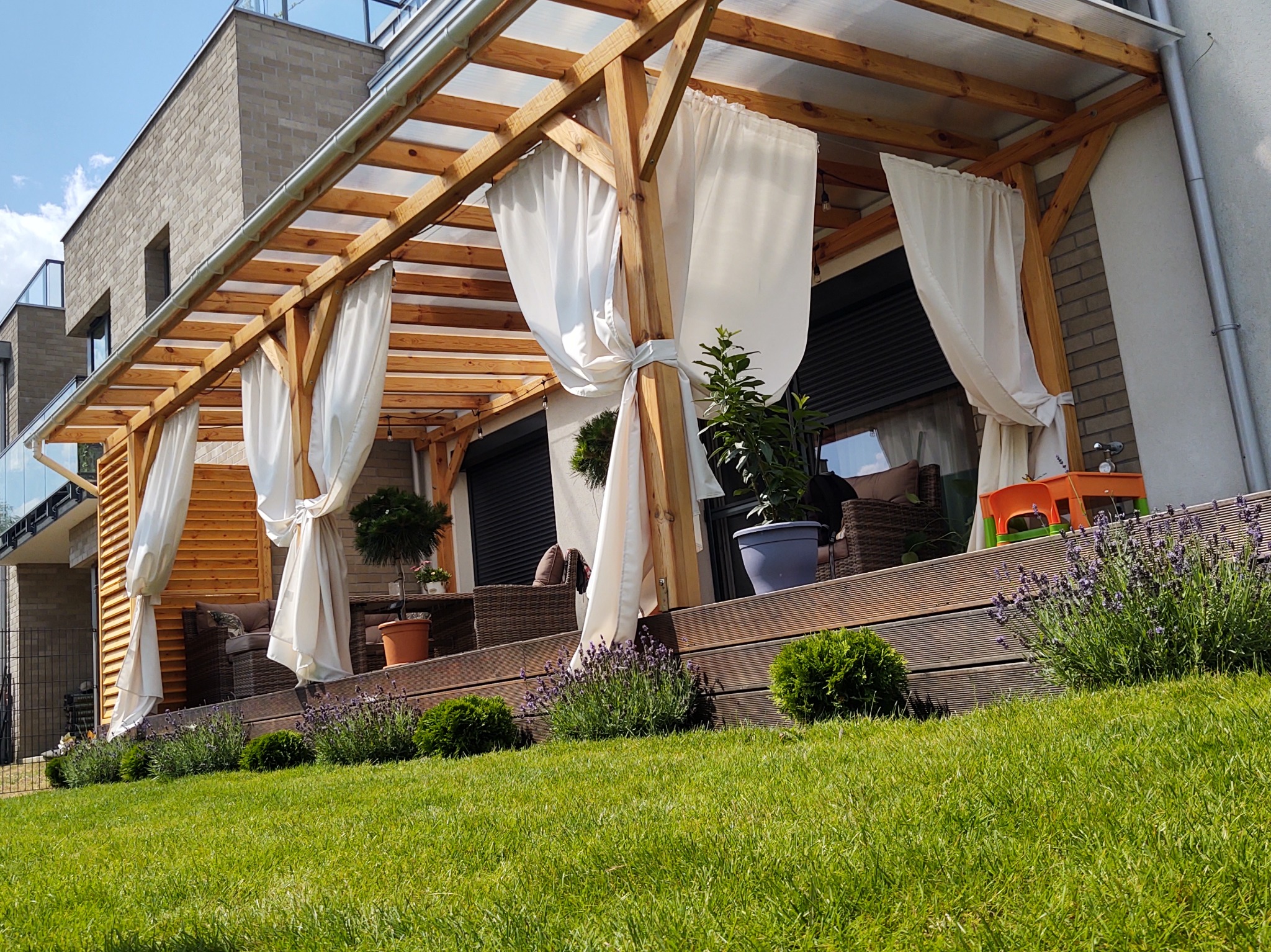 Drewniana pergola z białymi zasłonami i tarasem, otoczona zielonym trawnikiem i roślinami. Widoczne meble ogrodowe i elementy dekoracyjne.