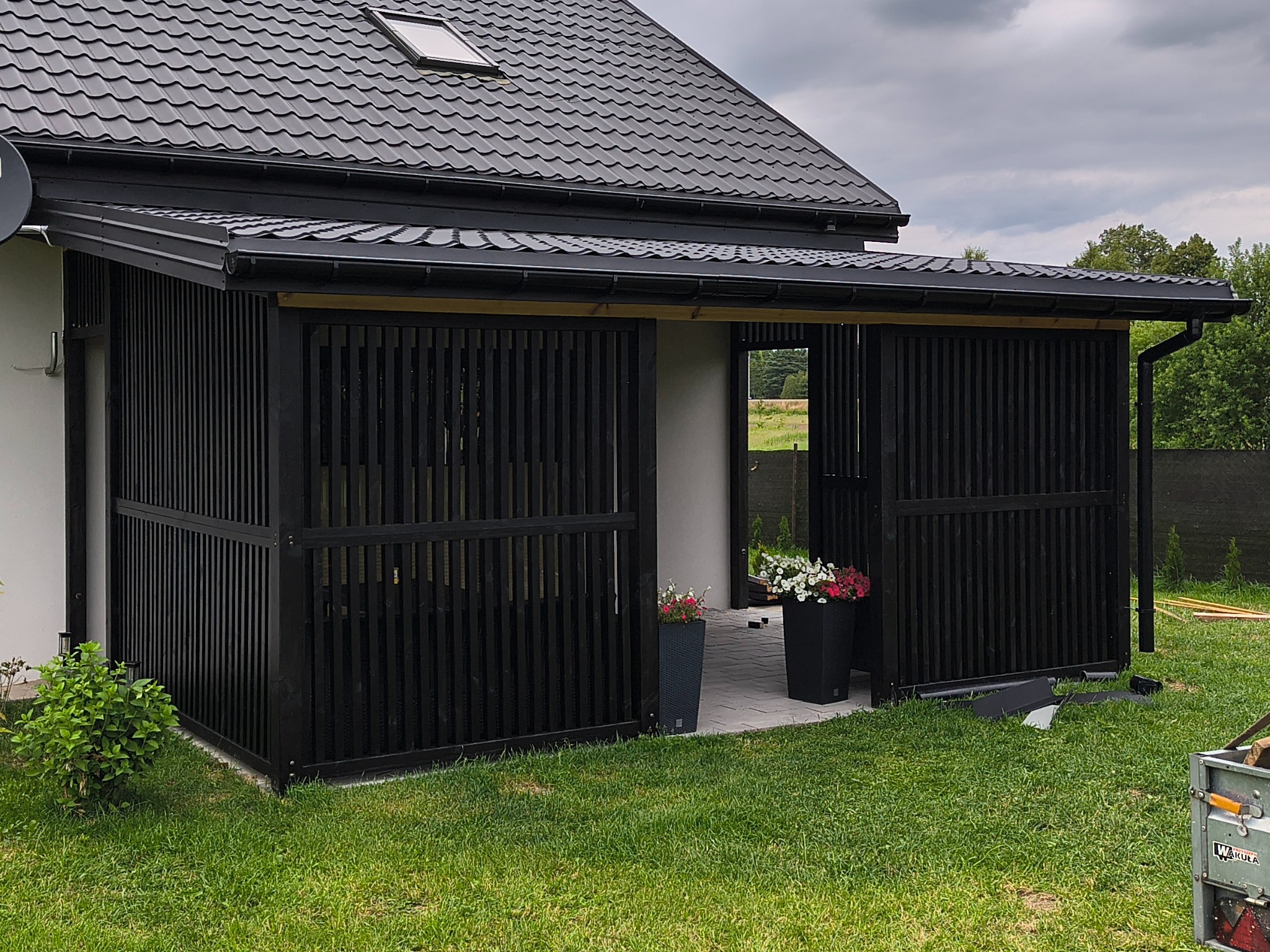 Nowoczesna, czarna pergola drewniana z pionowymi listwami, przylegająca do domu z ciemnym dachem. Widoczne donice z kwiatami i narzędzia na trawniku.