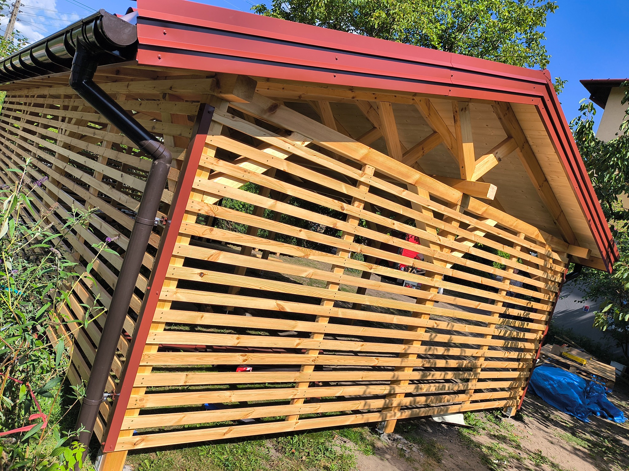 Drewniana konstrukcja z czerwonym dachem i brązową rynną, prawdopodobnie altana lub pergola, w trakcie budowy, z widocznymi elementami konstrukcyjnymi.
