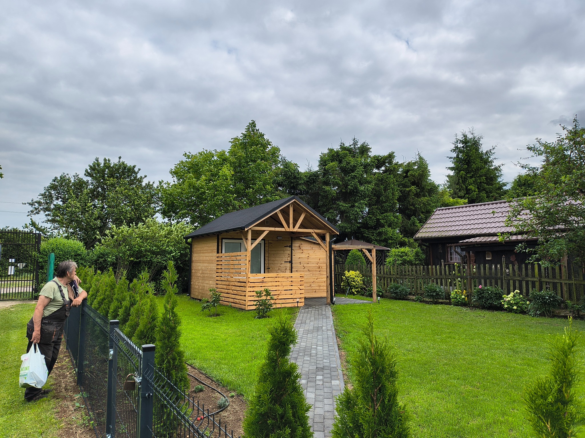 Drewniana altana z zadaszeniem i ścieżką z kostki w Grębkowie, otoczona zielenią. Osoba opiera się o ogrodzenie w lewym dolnym rogu. Pochmurne niebo w tle.