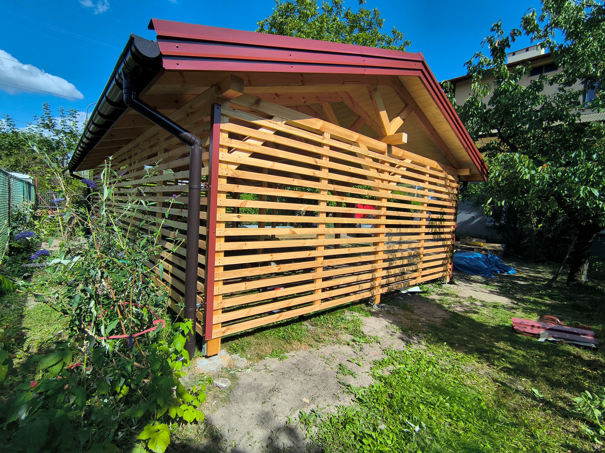 Nowa, drewniana pergola z brązowym dachem i poziomymi listwami, otoczona zielenią ogrodu w słoneczny dzień. Widoczne detale konstrukcyjne i elementy wykończeniowe.