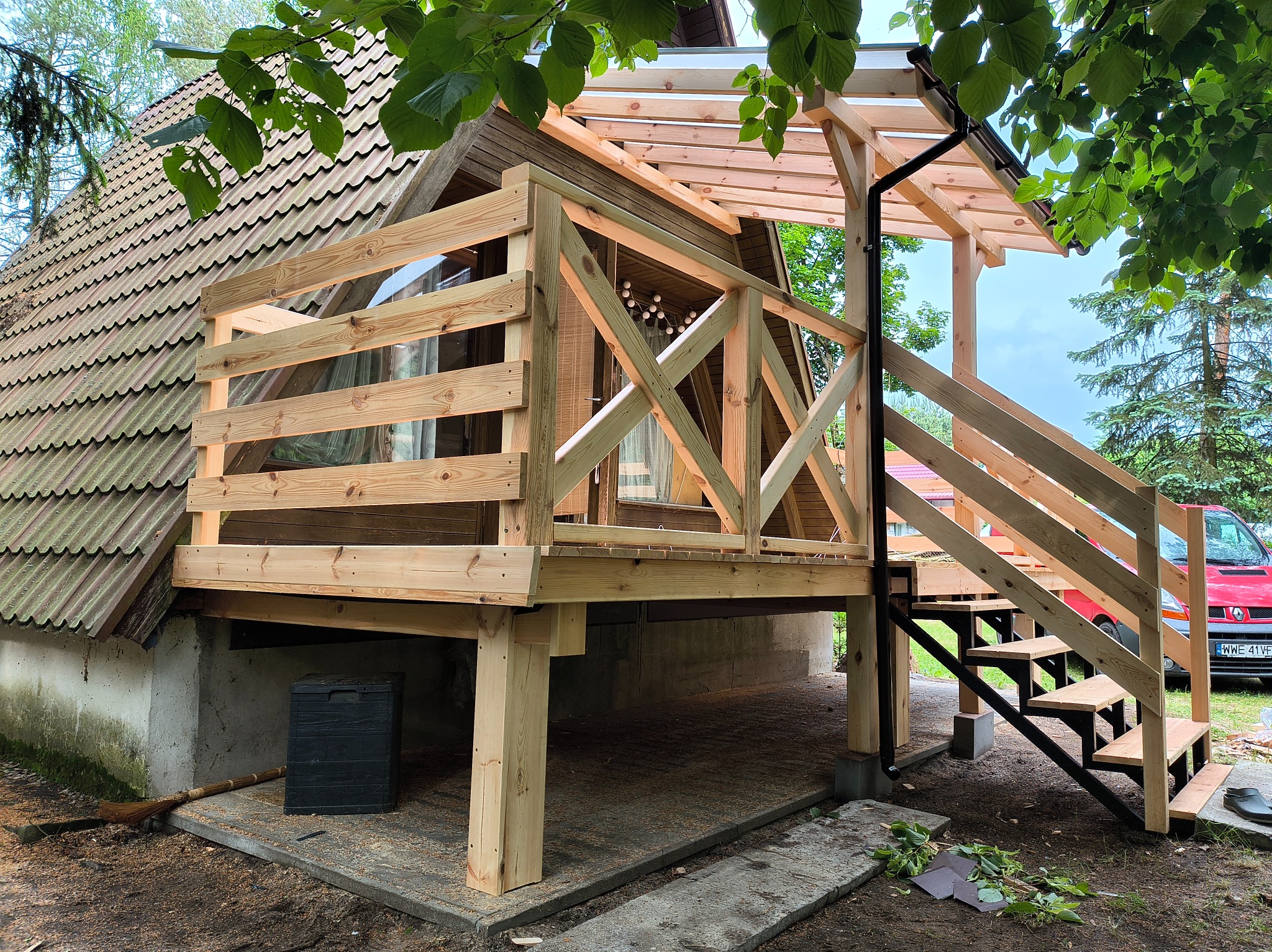 Nowa, drewniana konstrukcja tarasu z balustradą i schodami przy domu z dachem krytym dachówką. Widoczne elementy konstrukcyjne i fragment czerwonego samochodu w tle.