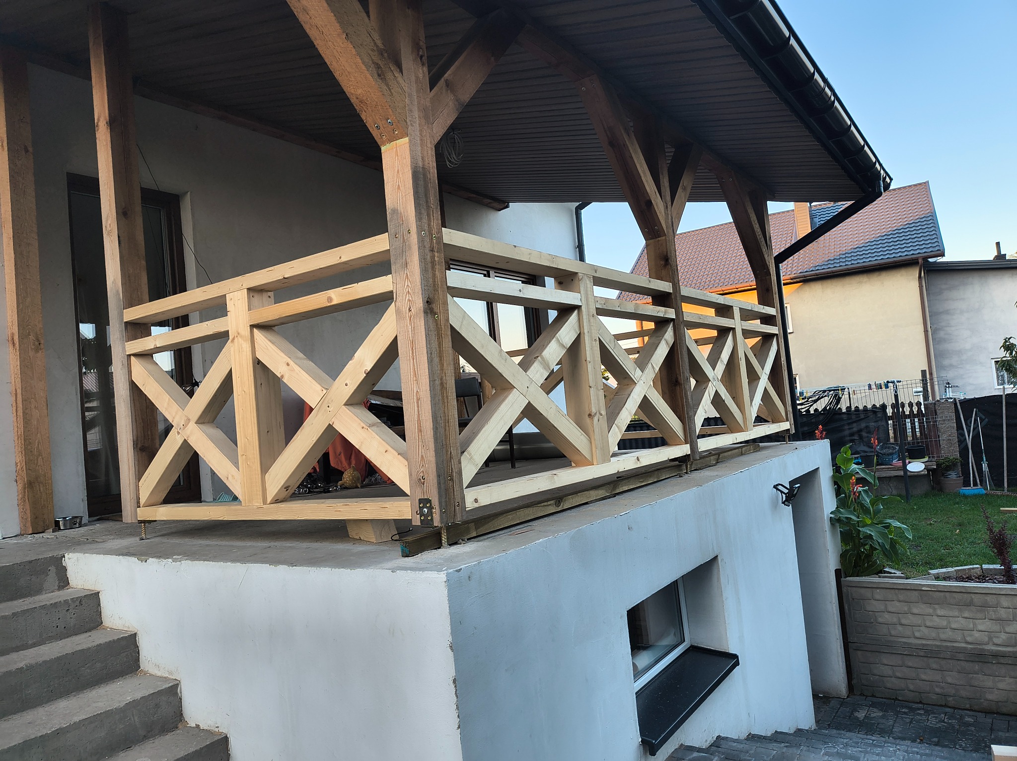 Drewniana balustrada z krzyżowym wzorem, świeżo po montażu, na tarasie z betonowymi schodami. Widoczne elementy konstrukcyjne i metalowe mocowania.