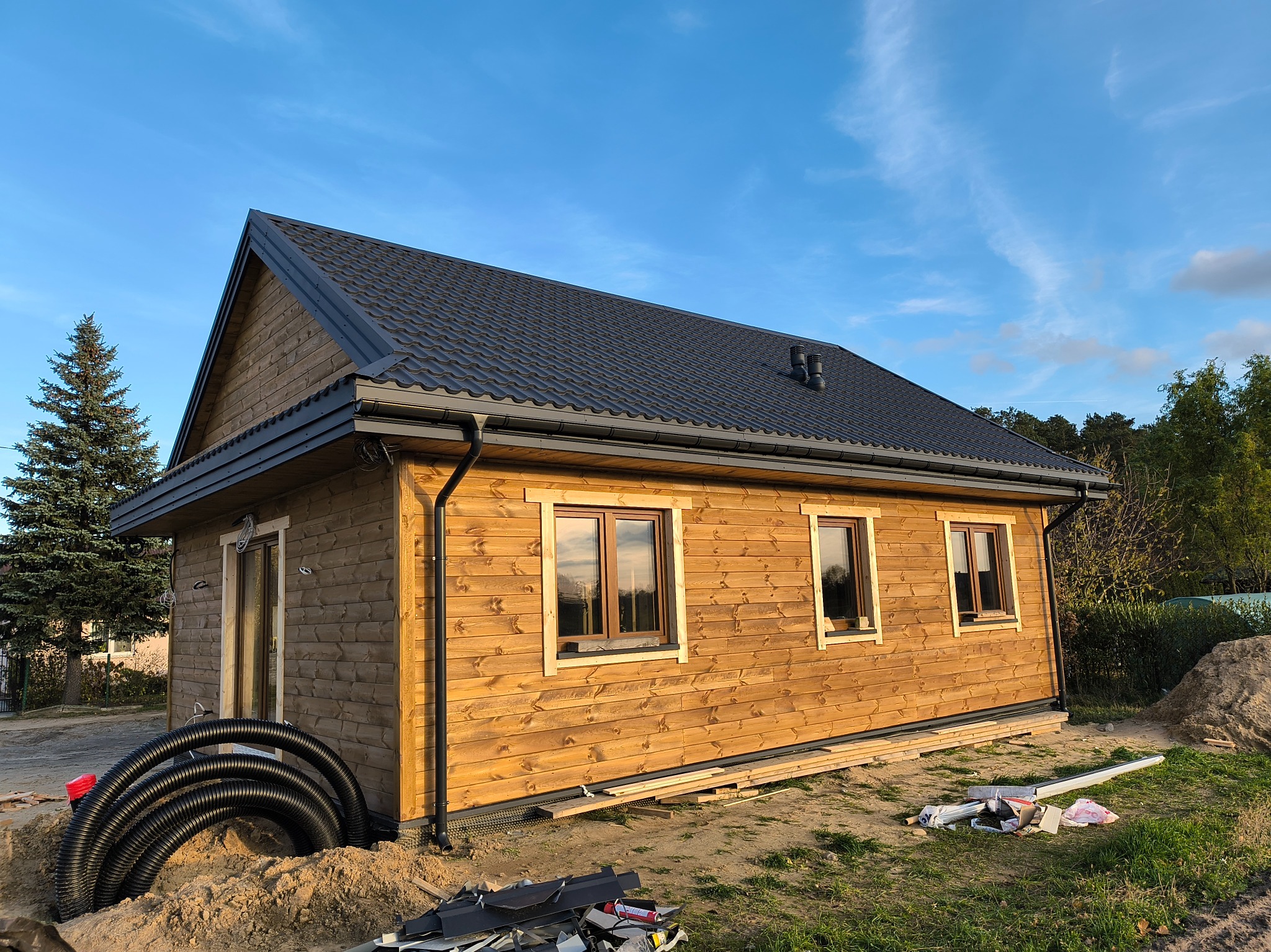 Drewniany dom szkieletowy z ciemnym dachem i widocznymi oknami, otoczony zielenią i elementami budowlanymi. Ujęcie w słoneczny dzień.