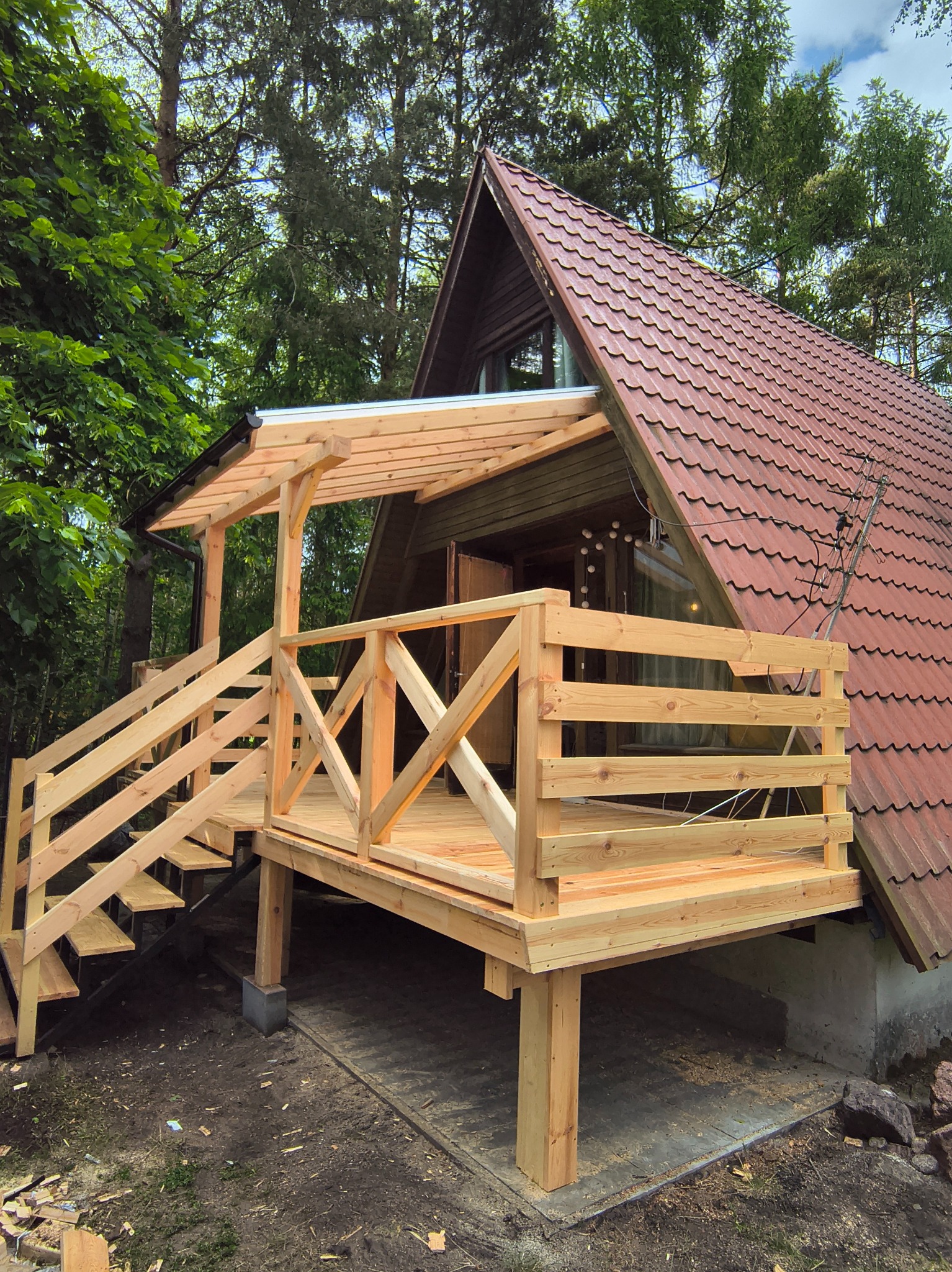 Nowy drewniany taras z zadaszeniem przy domu z czerwoną dachówką, otoczony zielenią. Widoczne schody i balustrady. Świeże drewno, naturalne otoczenie.