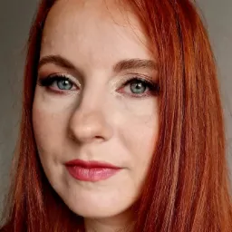 Portret kobiety z rudymi włosami i delikatnym makijażem oczu w odcieniach złota i różu, wykonanym w Gdyni, z uwydatnionymi rzęsami i pomalowanymi ustami na kolor malinowy.