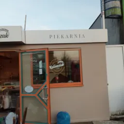 Świeżo otynkowana elewacja budynku piekarni z otwartymi drzwiami, widoczne narzędzia i pozostałości po pracach tynkarskich na chodniku.
