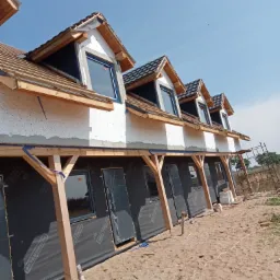 Budynek w trakcie ocieplania elewacji, widoczne okna dachowe, czarna membrana na ścianach i rusztowanie w tle.