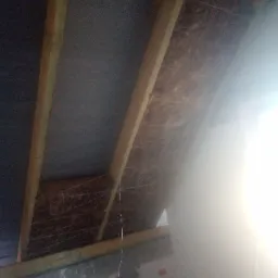 Skośny sufit poddasza z widoczną izolacją z wełny mineralnej, zabezpieczoną sznurkiem, pomiędzy drewnianymi belkami konstrukcyjnymi; fragment oświetlonej ściany w tle.