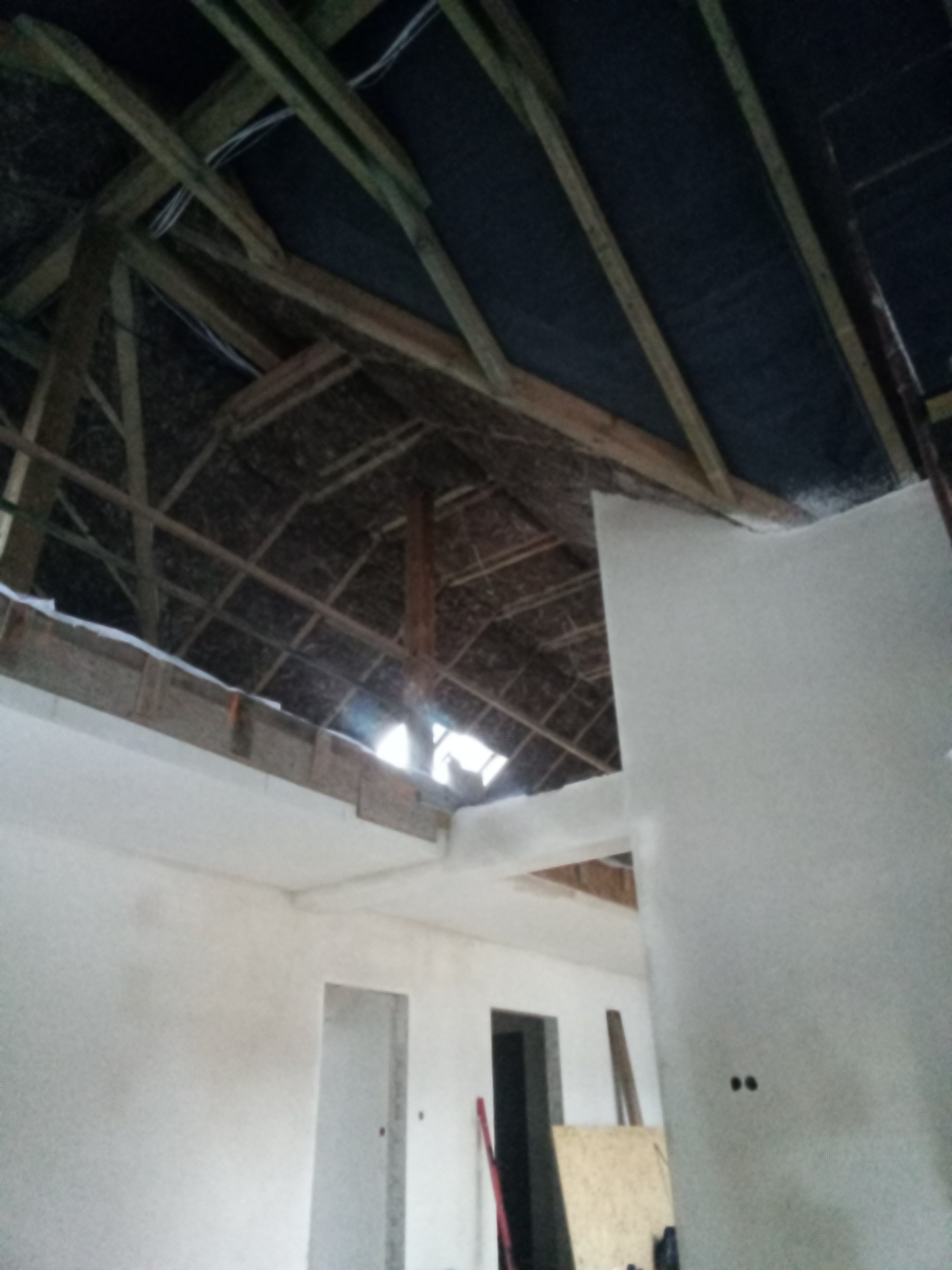 Surowe wnętrze poddasza z widoczną konstrukcją dachu, izolacją i fragmentem otynkowanej ściany, ukazujące etap prac wykończeniowych.