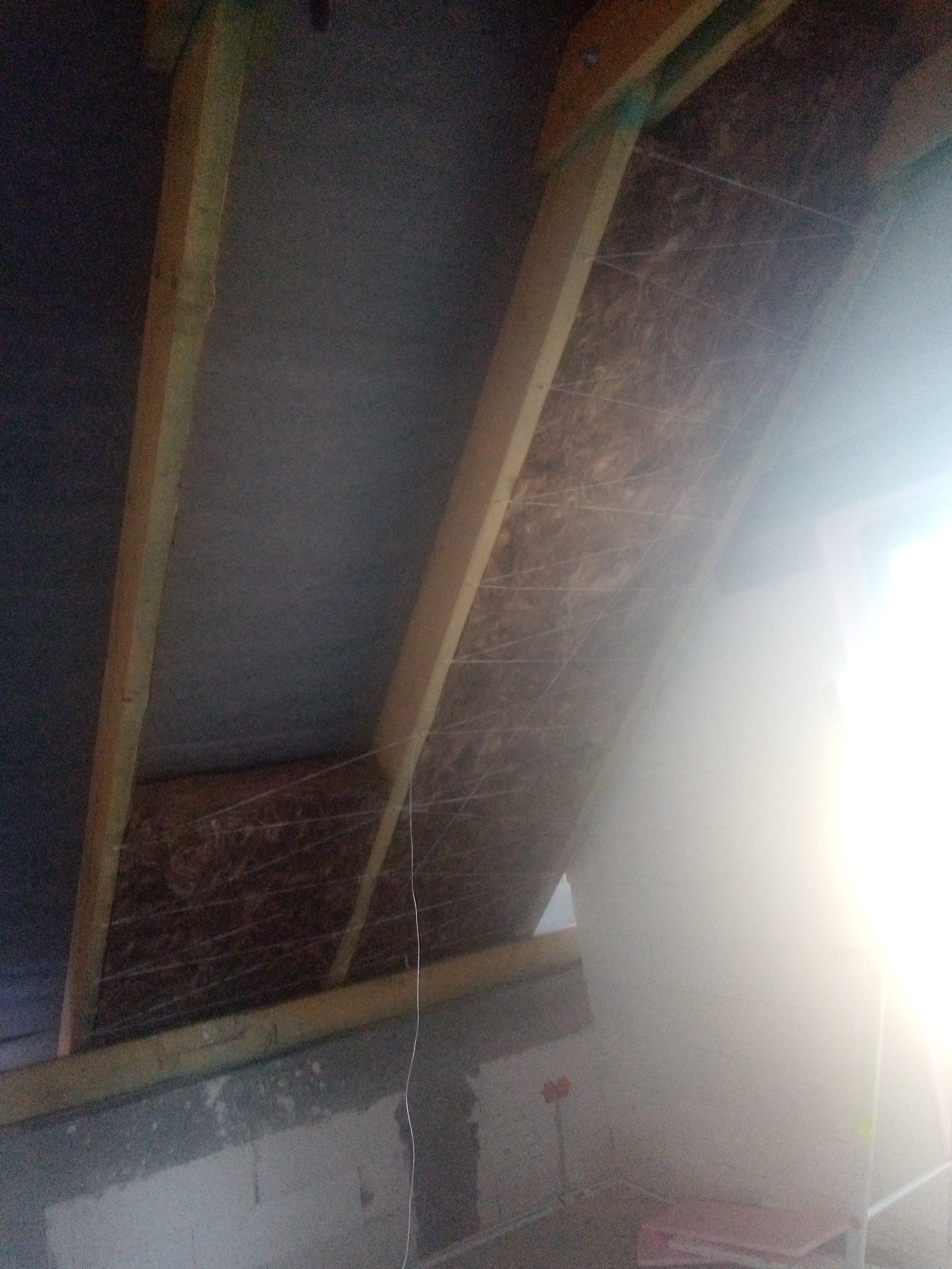 Skośny sufit poddasza z widoczną izolacją z wełny mineralnej, zabezpieczoną sznurkiem, pomiędzy drewnianymi belkami konstrukcyjnymi; fragment oświetlonej ściany w tle.